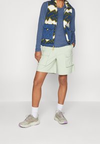 Osoba v modré košili s dlouhým rukávem, vzorované modro-bílé vestě s velkými kapsami, světle zelených cargo šortkách, bílých ponožkách a béžových teniskách s fialovými podrážkami.