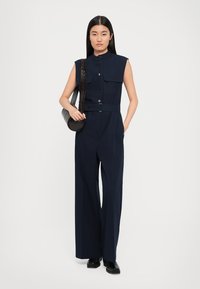 Navy sleeveless jumpsuit with a buttoned collar, two chest pockets, belted waist, and wide-leg design. Black round handbag and ankle boots.