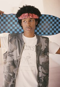 Patineta a cuadros negros y azules sostenida detrás de un joven que lleva un chaleco camuflado gris sobre una camiseta gráfica blanca con texto e imagen.