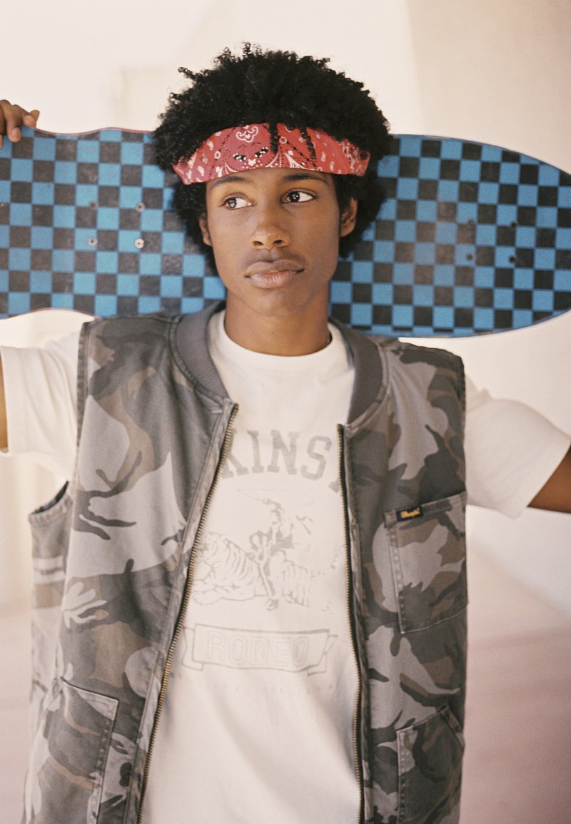 Patineta a cuadros negros y azules sostenida detrás de un joven que lleva un chaleco camuflado gris sobre una camiseta gráfica blanca con texto e imagen.