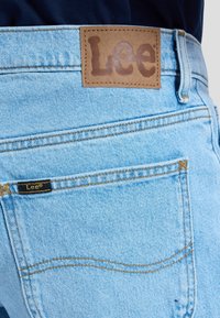 Tasca posteriore di jeans in denim azzurro chiaro con patch in pelle color cuoio del marchio Lee e piccola etichetta nera Lee sul bordo della tasca.