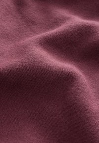 Tessuto ricco e strutturato in una profonda tonalità bordeaux, con pieghe morbide che creano delicati giochi di ombre e luci, esaltando la qualità del materiale.