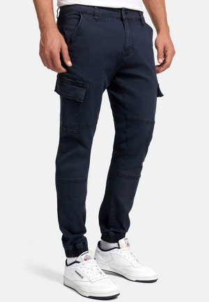Homme portant un pantalon cargo jogger bleu marine avec des poignets élastiques et des baskets blanches Reebok, debout devant un fond uni.