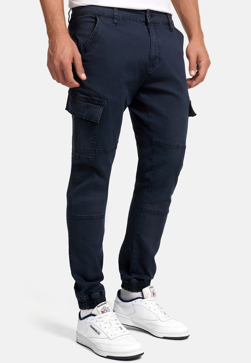 Homme portant un pantalon cargo jogger bleu marine avec des poignets élastiques et des baskets blanches Reebok, debout devant un fond uni.