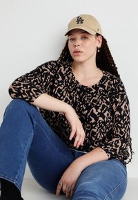 Långärmad topp i svart och beige leopardmönster, tillsammans med blå denimjeans. En beige keps med svart "LA"-logotyp bärs.