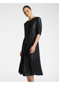 Schwarzes plissiertes Kleid aus einem glatten, glänzenden Stoff. Lange Ärmel, runder Halsausschnitt und fließender Rock verleihen eine entspannte Silhouette.