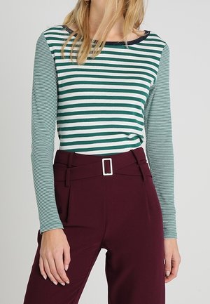 Femme portant un haut manches longues à rayures horizontales vertes et blanches avec un pantalon taille haute bordeaux avec une ceinture à boucle rectangulaire.