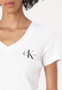 White cotton V-neck T-shirt featuring a black "CK" logo and "Calvin Klein Jeans" text on the left side of the chest. Short sleeves.