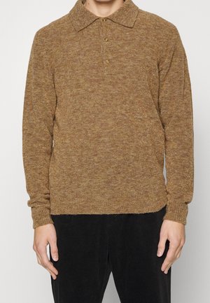 Persoon die een bruin gebreide polopullover met lange mouwen en drie knopen draagt, gecombineerd met een zwarte broek, staande tegen een witte achtergrond.