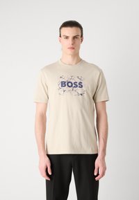 Beige bavlněné tričko s modrou grafickou potiskem na texturovaném pozadí. Kolaový výstřih a krátké rukávy, přiléhavý střih.