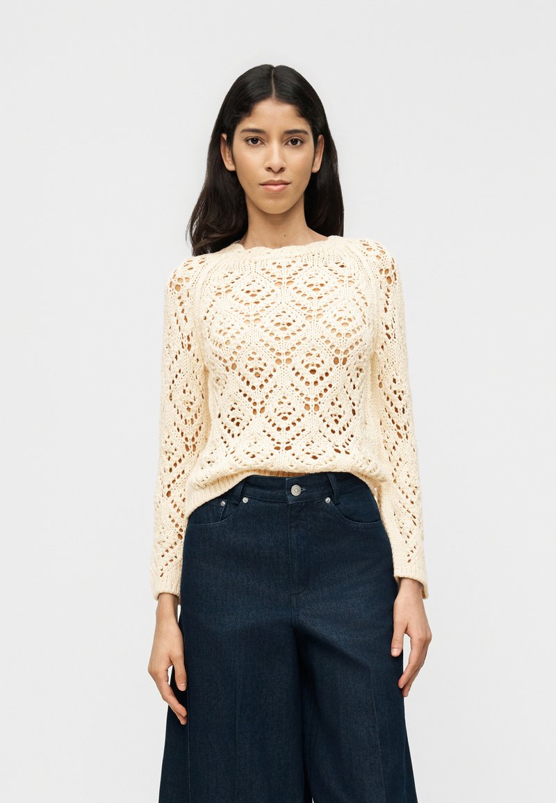 Νεαρή γυναίκα φορώντας μπεζ πλεκτό πουλόβερ και σκούρο μπλε τζιν με φαρδιά παντελόνια, στέκεται μπροστά σε απλό φωτεινό φόντο.