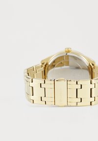 Montre en acier inoxydable doré avec un bracelet à maillons, boîtier rond, fond transparent et une couronne sur le côté. Logo gravé sur le fermoir.