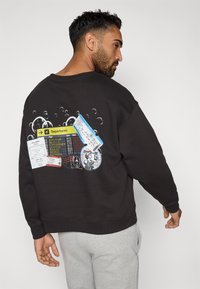 Melnas kapuces džemperis ar krāsainu grafisko dizainu uz muguras, kurā attēloti lidmašīnu biļetes, lidostas informācijas tablo un burbuļi.