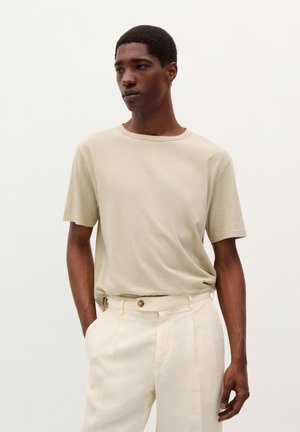 Beige Baumwoll-T-Shirt mit kurzen Ärmeln und rundem Halsausschnitt, kombiniert mit hellen, maßgeschneiderten Hosen mit einer vorderen Falte und Knopfdetails.