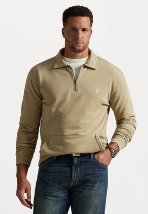 Man in een beige kwart-rits sweatshirt met een wit t-shirt eronder, blauwe jeans en een horloge om zijn link pols.