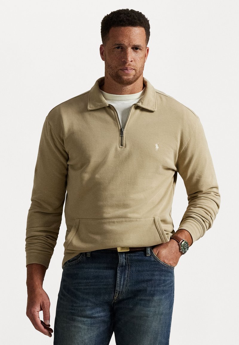 Un homme portant un sweat-shirt beige à zip quart avec un t-shirt blanc en dessous, un jean bleu et une montre au poignet gauche.