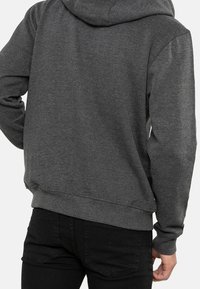 Vue de dos d'une personne portant un sweat à capuche gris foncé et un jean noir, les mains détendues le long du corps.