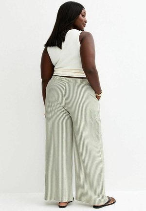 Mujer con cabello largo y oscuro, vistiendo una blusa blanca sin mangas y pantalones anchos a rayas, posando frente a un fondo blanco.