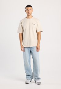 Beige puuvillane oversize t-särk, ümmarguse kaelusega, lühikeste varrukatega ja väikese tikitud logoga. Koos hallikas sinise denimist teksapükstega ja spordijalatsitega.