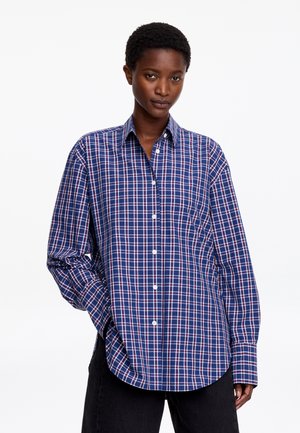 Persona que lleva una camisa de cuadros azul, blanca y roja con botones y mangas largas, de pie con una mano en el bolsillo sobre un fondo blanco.