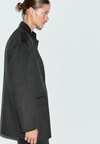 Cappotto oversize grigio scuro con colletto alto, chiusura a bottoni e tasche laterali; realizzato in tessuto morbido con una texture liscia.
