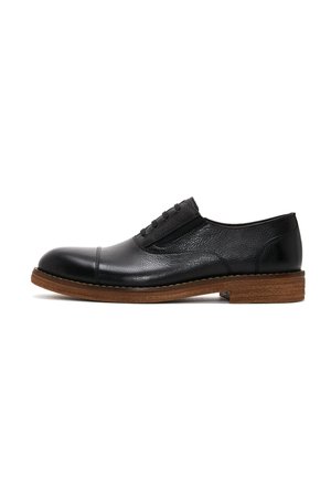 Zapato negro de cuero con cordones, con una punta lisa y un cuerpo texturizado. Presenta paneles elásticos y una suela de goma de color marrón claro.