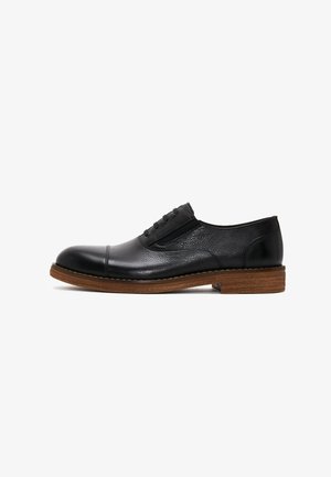 Chaussure noire en cuir à lacets avec un bout lisse et un corps texturé. Présente des panneaux élastiques et une semelle en caoutchouc beige clair.