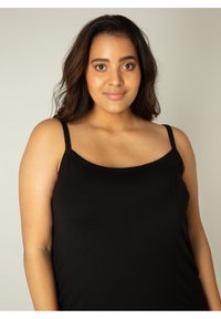 Jonge vrouw met halflang donker haar, gekleed in een zwarte mouwloze top, staand tegen een eenvoudige lichte achtergrond, zacht glimlachend.