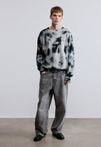 Μπλούζα μπλουζάκι μαύρου και γκρι tie-dye με ριμπ μανσέτες, ταμπέλα λογότυπου στο στήθος, συνδυασμένη με φαρδιά γκρι τζιν τζιν και μαύρα παπούτσια slip-on.