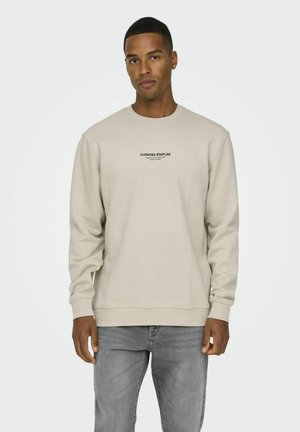 Beige crewneck sweatshirt med ribbede manchetter og kant, med et lille sort "CURATED STAPLES" logo i midten foran. Grå jeans.