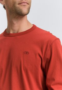 Rotes Langarmshirt aus glattem Stoff, mit Rundhalsausschnitt, verstärkten Nähten und kleinem Logo auf der Brust.