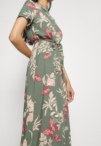 Abito floreale verde con maniche corte, caratterizzato da una chiusura con bottoni e motivi di farfalle rosa. Vita con cintura e silhouette fluida.