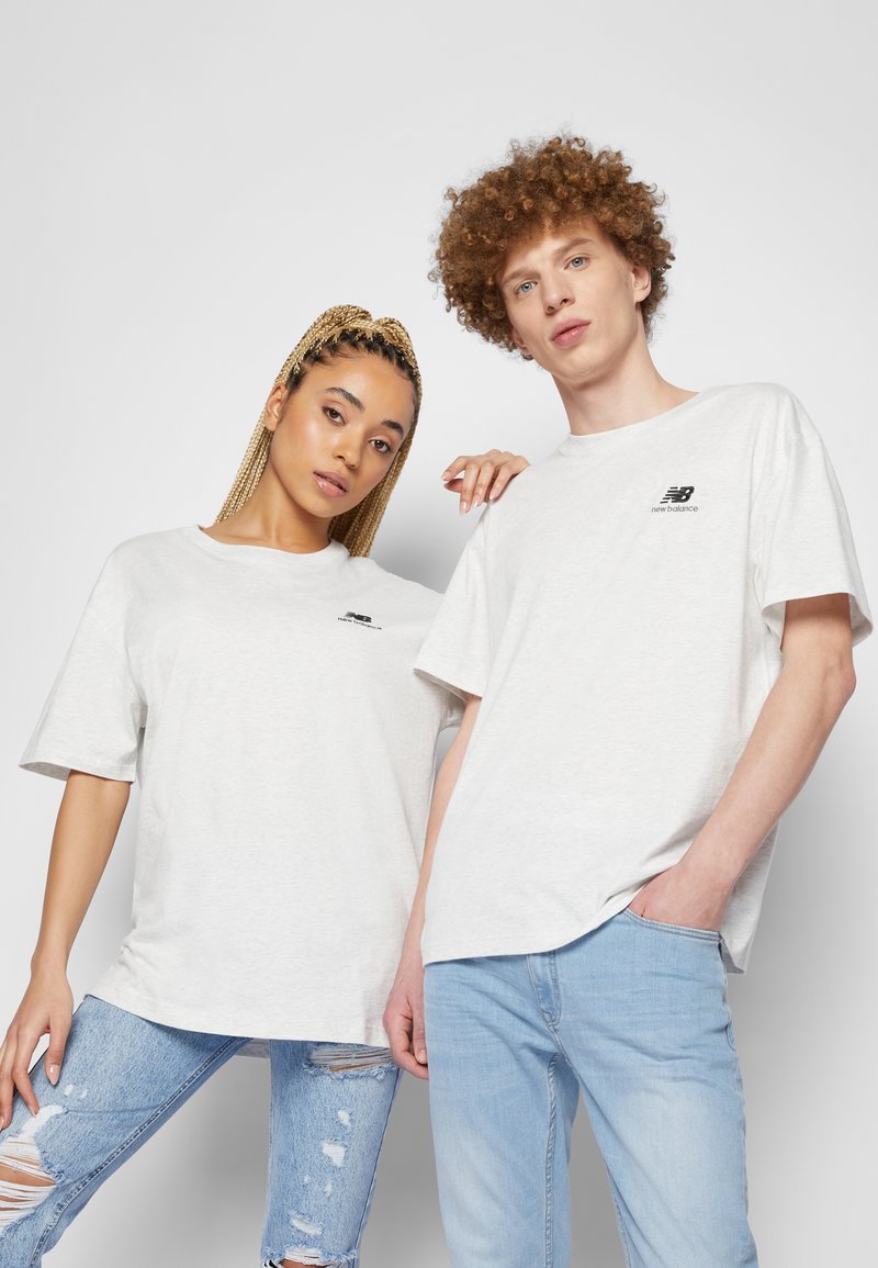 Due modelli indossano T-shirt grigie chiare di New Balance e jeans strappati di un blu chiaro, posando contro uno sfondo grigio semplice.