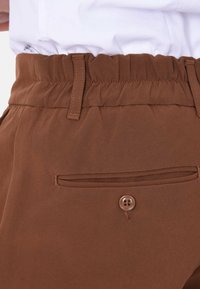 Pantaloni in tessuto marrone con vita elasticizzata, tasche laterali e una tasca a filetto sul retro con un dettaglio in scatto marrone. Texture liscia.