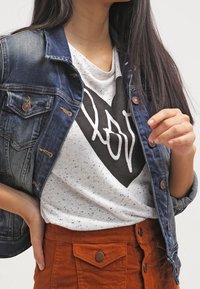 Chaqueta de mezclilla con botones de bronce, combinada con una camiseta blanca con manchas que presenta un atrevido diseño de corazón negro y la palabra "love", junto con una falda de pana de color óxido.