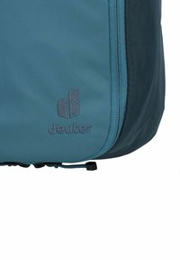 Mochila de color verde azulado que cuenta con una tela suave y duradera. Incluye un zipper con una tira de tirón azul y un acento azul oscuro cerca de la base. Logotipo de Deuter visible.