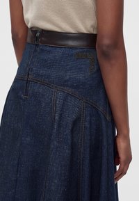 Gros plan d'une personne portant une jupe en jean bleu foncé avec des surpiqûres marron et une ceinture en cuir noire, montrant partiellement un logo "MAX & Co".