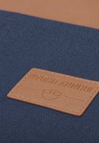 Plátno v tmavě modré barvě s texturovaným povrchem; na jedné straně se nachází béžová kožená nášivka s vyraženým brandingem.