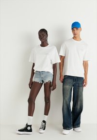 Valged T-särgid sisaldavad trükitud detaile. Ühel inimesel on seljas teksashorts ja mustad sneakerid; teisel on seljas laiad teksad ja sinine müts.