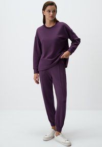 Paarse sweatshirt met zijsplits, gecombineerd met bijpassende taps toelopende sweatpants. De stof heeft een gladde textuur. Model draagt witte sneakers.