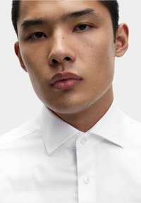 Junger Mann mit neutralem Gesichtsausdruck, der ein frisches weißes, geknöpftes Hemd vor einem schlichten hellen Hintergrund trägt.