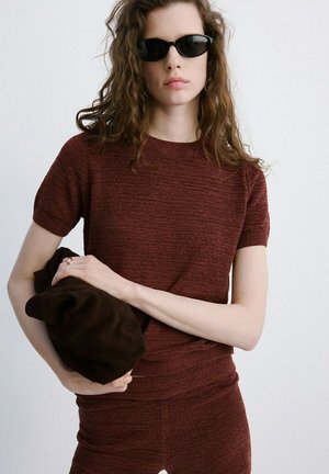 Femme aux cheveux bouclés portant des lunettes de soleil foncées et un haut à manches courtes ainsi que un pantalon assortis, tenant un accessoire ou sac doux brun foncé.