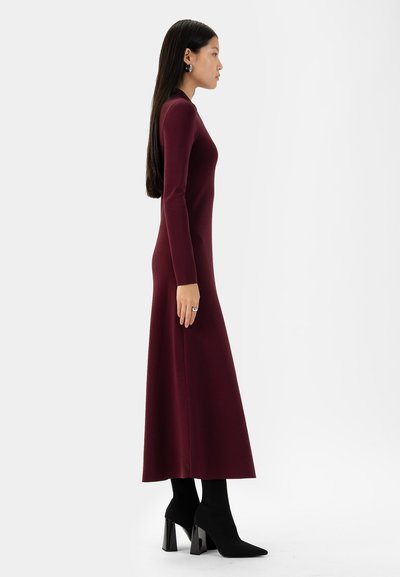 Lungo abito in maglia bordeaux con maniche lunghe, corsetto aderente e orlo svasato; abbinato a stivaletti neri con punta e a un semplice anello d'argento.