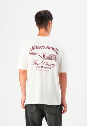 Man draagt een wit T-shirt met bordeauxrode tekst en vorkgrafiek, houdt spaghetti op zijn rug vast, gecombineerd met donkere spijkerbroek, met zijn gezicht van je af gewend.