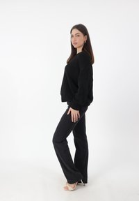 Zwarte oversized trui met geribde manchetten, gecombineerd met strakke zwarte jeans. Model staat zijwaarts, met beige hoge hakken.