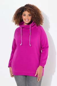 Lachende vrouw met krullend haar, gekleed in een felroze hoodie met witte trekkoorden en grijze broek tegen een effen achtergrond.