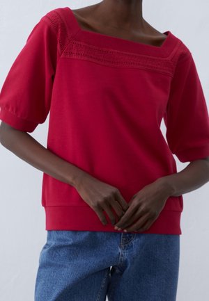Persoon draagt een rood topje met korte mouwen en een vierkante halslijn met kantdetail, en een blauwe spijkerbroek, handen losjes in elkaar gestrengeld op de taille.