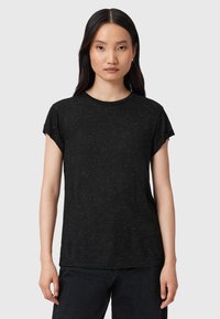 Mujer con cabello largo y oscuro, vestida con una camiseta negra con manchas y pantalones oscuros, de pie contra un fondo claro y liso.