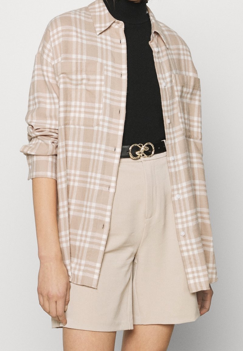 Femme portant une chemise beige à carreaux sur un col roulé noir, un short beige et une ceinture noire avec une boucle double anneau dorée.