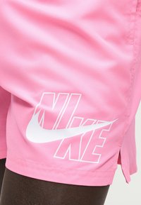 Ροζ αθλητικό σορτς με λευκό λογότυπο Nike τυπωμένο κοντά στο τελείωμα σε άτομο με σκουρόχρωμο δέρμα.